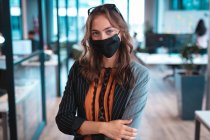 Portrait d'une femme d'affaires caucasienne portant un masque facial avec des collègues en arrière-plan. travailler dans un bureau moderne pendant la pandémie de coronavirus covid 19. — Photo de stock