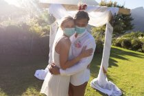 Felici sposi caucasici che si sposano indossando maschere facciali e abbracciando. matrimonio estivo, matrimonio, amore e celebrazione durante covid 19 concetto pandemico. — Foto stock
