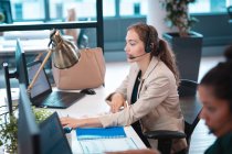 Donna d'affari caucasica con le cuffie e seduta a tavola a parlare, usando il computer. lavorare in un ufficio moderno. — Foto stock