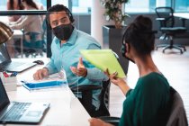 Dos empresarios diversos que usan mascarilla facial, que tienen documentos y utilizan la computadora. trabajar en una oficina moderna durante la pandemia de coronavirus covid 19. - foto de stock