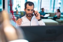 Gemischte Rasse Geschäftsmann sitzt am Tisch mit Smartphone mit Kollegen im Hintergrund. Arbeit in einem modernen Büro. — Stockfoto