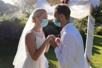 Happy caucasian bride and groom getting married wearing face masks and holding hands. summer wedding, marriage, love and celebration during covid 19 pandemic concept. — Stock Photo