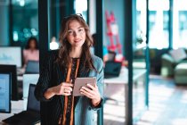 Retrato de mujer de negocios caucásica sonriendo y usando tableta con colegas de fondo. trabajar en una oficina moderna. - foto de stock