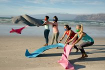 Un grupo de diversas amigas con máscaras en la cara que dejan alfombras, practican yoga, en la playa. estilo de vida activo saludable, fitness al aire libre y bienestar durante la pandemia de covid 19. - foto de stock