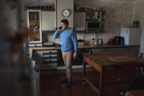 Focused caucasian man standing in kitchen, looking out window, using smartphone. spending free time at home. — Stock Photo