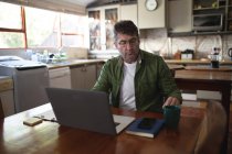 Uomo caucasico in cucina seduto a tavola usando laptop e scrivendo appunti. tecnologia e comunicazione, lavoro flessibile da casa. — Foto stock