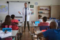 Кавказский врач-мужчина разговаривает с группой студентов, сидящих в классе в школе. охрана здоровья и безопасность в школе во время пандемии ковид-19 — стоковое фото