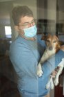 Porträt eines kaukasischen Mannes mit Brille und Gesichtsmaske, Hund haltend, durch Fenster schauend. Zeit zu Hause verbringen, während Coronavirus covid 19 Pandemie. — Stockfoto