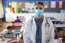 Retrato de médico caucasiano vestindo máscara facial em pé na classe na escola. protecção da saúde e segurança na escola durante o conceito de pandemia de covid-19 — Fotografia de Stock