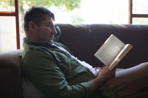 Homem caucasiano sorridente deitado no sofá, lendo livro e relaxante. passar tempo livre em casa. — Fotografia de Stock