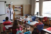 Kaukasischer Arzt im Gespräch mit einer Gruppe verschiedener Schüler, die in der Schulklasse sitzen. Gesundheitsschutz und Sicherheit in der Schule während des Covid-19-Pandemiekonzepts — Stockfoto