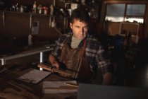 Caucasian male knife maker holding knife, using laptop in workshop. independent small business craftsman at work. — Stock Photo