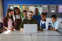 Insegnante maschio caucasico e gruppo di studenti diversi che utilizzano il computer portatile insieme nella classe a scuola. concetto di scuola e istruzione — Foto stock