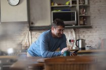 Улыбающийся кавказский мужчина на кухне, стоящий за столом, пьющий кофе и пользующийся смартфоном. проводить свободное время дома. — стоковое фото