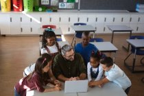 Учитель-кавказский мужчина и группа различных учеников вместе используют ноутбук в классе в школе. школа и концепция образования — стоковое фото