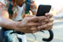Afrikanischer Mann in der Stadt sitzt auf dem Fahrrad und benutzt ein Smartphone. digitaler Nomade unterwegs, unterwegs in der Stadt. — Stockfoto