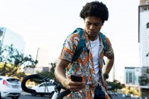 Homem afro-americano na cidade sentado de bicicleta e usando smartphone. nômade digital em movimento, para fora e sobre na cidade. — Fotografia de Stock