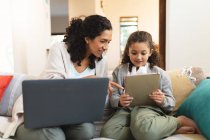 Sorridente razza mista madre e figlia seduta sul divano, utilizzando laptop e tablet. stile di vita domestico e trascorrere del tempo di qualità a casa. — Foto stock