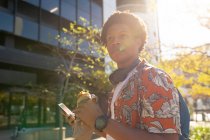 Hombre afroamericano en la ciudad comiendo y usando smartphone. nómada digital sobre la marcha, fuera y alrededor de la ciudad. - foto de stock