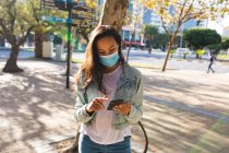 Азійка, одягнена в маску обличчя, користується смартфоном у сонячному парку. Незалежна молода жінка, яка живе в місті під час коронавірусу (19 пандемії). — стокове фото