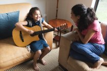 Raza mixta madre e hija sentadas en el sofá y tocando la guitarra. estilo de vida doméstico y pasar tiempo de calidad en casa. - foto de stock