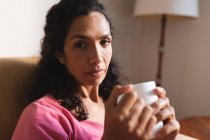 Portrait of mixed race woman sitting on sofa and drinking coffee. domestic lifestyle and spending quality time at home. — Stock Photo