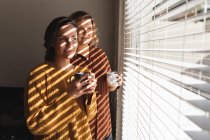 Лесбійська пара посміхається і п'є каву поруч з вікном. Домашнє життя, вільний час удома. — стокове фото