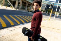 Uomo afroamericano in citta 'che tiene lo skateboard. nomade digitale in movimento, in giro per la città. — Foto stock
