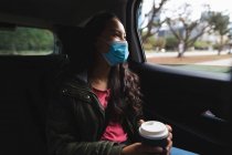 Asiatin mit Gesichtsmaske sitzt im Taxi und hält Kaffee zum Mitnehmen. Unabhängige junge Frau während der Coronavirus-Pandemie 19 in der Stadt unterwegs. — Stockfoto
