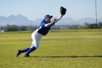 Смешанная раса женщина бейсболистка на солнечном бейсбольном поле тянется, чтобы поймать мяч во время игры. женская бейсбольная команда, спортивная подготовка и тактика игры. — стоковое фото