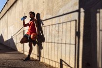 Ajuste hombre afroamericano ejercicio en la ciudad celebración de baloncesto y agua potable en la calle. fitness y estilo de vida urbano activo al aire libre. - foto de stock