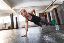 Mujer caucásica fuerte haciendo ejercicio en el gimnasio, haciendo flexiones con una mano. entrenamiento cruzado de fuerza y fitness para boxeo. - foto de stock