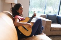 Mujer de raza mixta sentada en el sofá y tocando la guitarra. estilo de vida doméstico y pasar tiempo de calidad en casa. - foto de stock