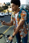 Homme afro-américain en ville assis sur un vélo et en utilisant un smartphone. nomade numérique en déplacement, en déplacement dans la ville. — Photo de stock