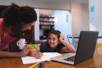 Sorridente madre di razza mista che tiene il caffè, aiutando sua figlia a fare i compiti. stile di vita domestico e trascorrere del tempo di qualità a casa. — Foto stock