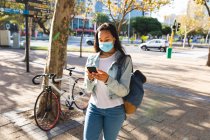 Mujer asiática que usa mascarilla facial usando smartphone en Sunny Park. mujer joven independiente fuera y alrededor de la ciudad durante coronavirus covid 19 pandemia. - foto de stock