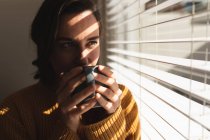 Donna caucasica sorride e beve caffè vicino alla finestra. stile di vita domestico, trascorrere il tempo libero a casa. — Foto stock