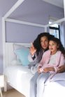 Feliz mestiço mãe e filha sentados na cama, abraçando e olhando através da janela. estilo de vida doméstico e passar tempo de qualidade em casa. — Fotografia de Stock