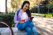 Усміхнена азіатська жінка носить навушники за допомогою смартфона і тримає каву на виході в сонячному парку. незалежна молода жінка на вулиці і близько в місті . — стокове фото
