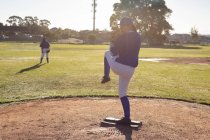 Lanzador de béisbol femenino de raza mixta en el campo de béisbol soleado preparándose para lanzar pelota durante el juego. equipo femenino de béisbol, entrenamiento deportivo y tácticas de juego. - foto de stock