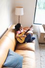 Mujer de raza mixta sentada en el sofá y tocando la guitarra. estilo de vida doméstico y pasar tiempo de calidad en casa. - foto de stock