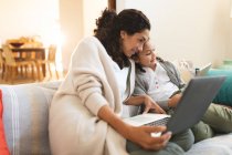 Sorrindo mestiço mãe e filha sentados no sofá, usando laptop e tablet. estilo de vida doméstico e passar tempo de qualidade em casa. — Fotografia de Stock