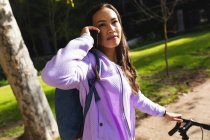 Портрет улыбающейся азиатки, катающейся на велосипеде и разговаривающей на смартфоне в солнечном парке. независимая молодая женщина в городе. — стоковое фото