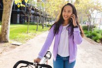 Mujer asiática sonriente rodando bicicleta y hablando en el teléfono inteligente en el parque soleado. mujer joven independiente fuera y alrededor de la ciudad. - foto de stock