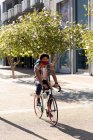 African american man in city wearing face mask cycling. digital nomad on the go, out and about in the city during coronavirus covid 19 pandemic. — Stock Photo