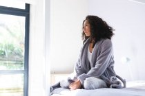 Счастливая задумчивая расовая женщина сидит на кровати и смотрит в окно. домашний образ жизни и проводить время дома. — стоковое фото
