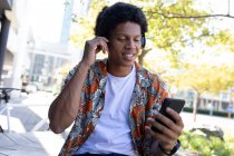 Uomo afroamericano in città seduto e utilizzando smartphone. nomade digitale in movimento, in giro per la città. — Foto stock