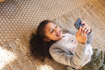 Sorrindo menina de raça mista deitado no tapete usando smartphone. estilo de vida doméstico e passar tempo de qualidade em casa. — Fotografia de Stock