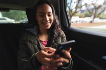 Lächelnde Asiatin, die im Taxi sitzt und ihr Smartphone benutzt. Unabhängige junge Frau in der Stadt unterwegs. — Stockfoto