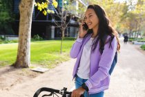 Sorrindo mulher asiática de bicicleta de rodas e falando no smartphone no parque ensolarado. jovem independente para fora e sobre na cidade. — Fotografia de Stock